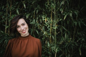 Sophie, debout la tête sur le coté, sourriante. Elle porte une blouse marron,  devant des bambous